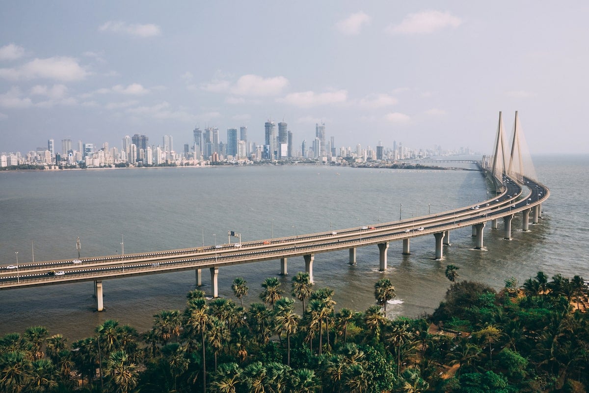 Aerial view of the Bandra-Worli Sea Link in Mumbai, a stunning cable-stayed bridge stretching over the Arabian Sea, with the city's modern skyline in the background and lush greenery in the foreground.