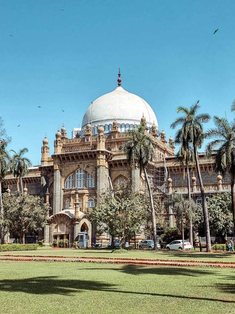 Exterior view of the Chhatrapati Shivaji Maharaj Vastu Sangrahalaya (formerly Prince of Wales Museum) in Mumbai, showcasing its grand Indo-Saracenic architecture with a prominent white dome, ornate arches, and lush palm trees in the foreground.