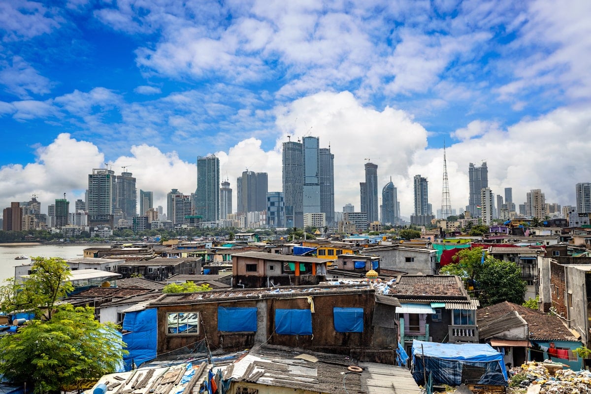 Contrasting view of Dharavi in Mumbai, with its dense informal housing in the foreground and the modern city skyline of high-rise buildings under a vibrant blue sky in the background.