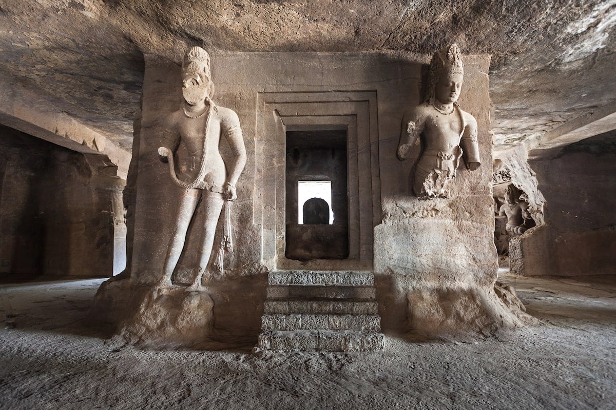 Intricately carved stone sculptures inside the Elephanta Caves in Mumbai, showcasing ancient Hindu deities and temple architecture, with a central doorway leading further into the historic rock-cut complex.