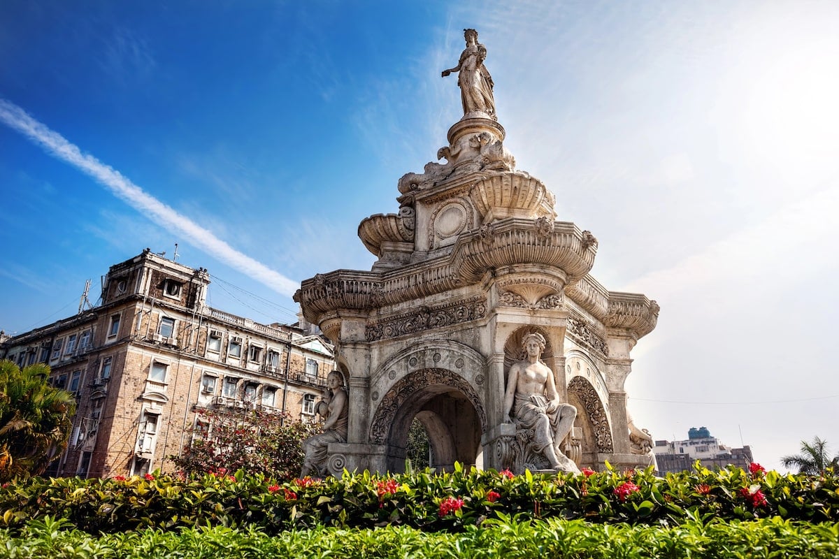 Flora Fountain in Mumbai, a beautifully carved heritage monument featuring intricate sculptures and a statue of the Roman goddess Flora, surrounded by lush greenery and set against a bright blue sky.