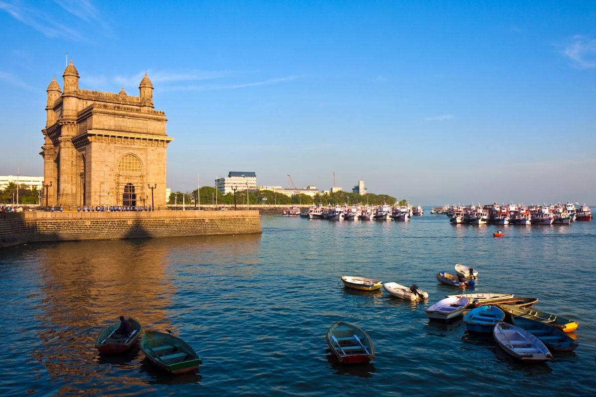 The Gateway of India in Mumbai, a grand arch monument and popular attraction on the waterfront, with colorful boats anchored in the Arabian Sea and a clear blue sky in the background.