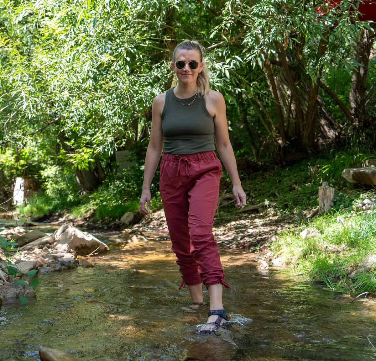 female hiker wearing Coalatree's Trailhead Pants in a rust color while crossing over a stream