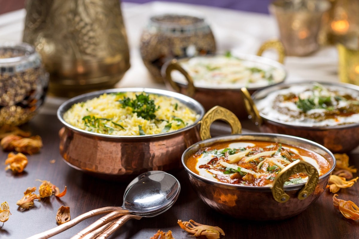 Solo female traveler in Mumbai sitting at a table of traditional Indian dishes served in copper bowls, including fragrant rice and creamy curry, beautifully garnished and accompanied by intricate decor in the background.