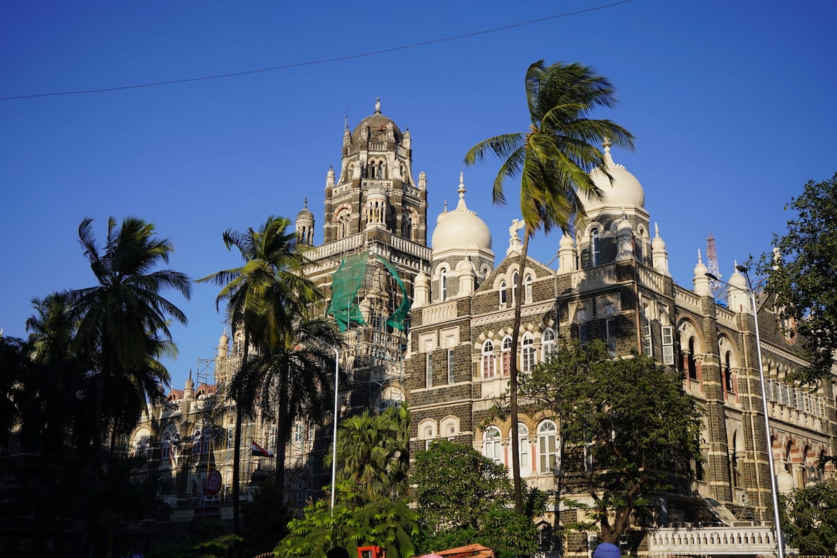 Detailed view of the historic Indo-Saracenic architecture of the Western Railway Headquarters building in the Fort district of Mumbai, India, featuring grand stone towers, white domes, and scaffolding for restoration, surrounded by tall green palm trees under a clear blue sky.
