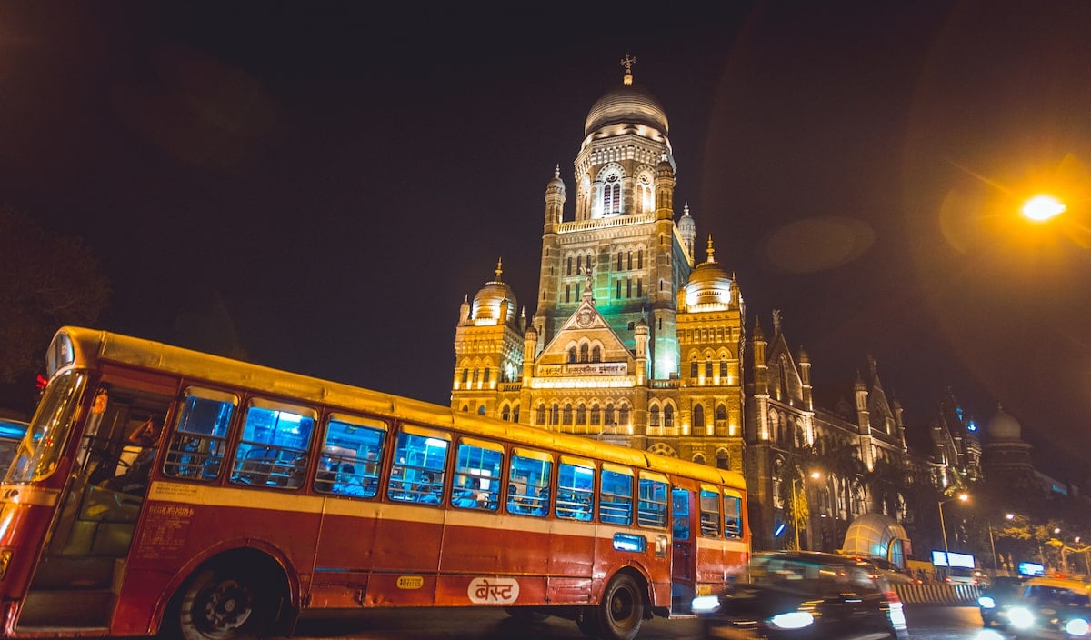 Night view of Mumbai with a bright red city bus in the foreground and the illuminated BMC (Brihanmumbai Municipal Corporation) building showcasing its striking colonial architecture in the background.