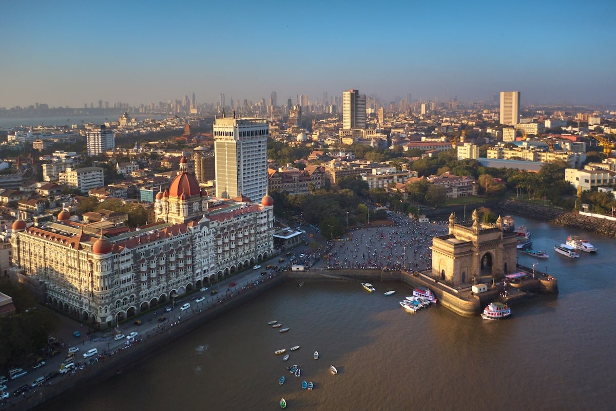 Aerial view of the Mumbai skyline featuring the iconic Gateway of India and the Taj Mahal Palace hotel on the waterfront, with boats in the harbor and the cityscape extending into the background under a clear blue sky.