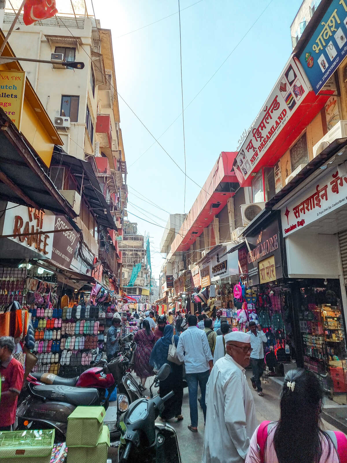 Busy street scene at Crawford Market in Mumbai, lined with colorful stalls selling textiles, bags, and other goods, with shoppers and motorbikes navigating the vibrant, bustling marketplace.