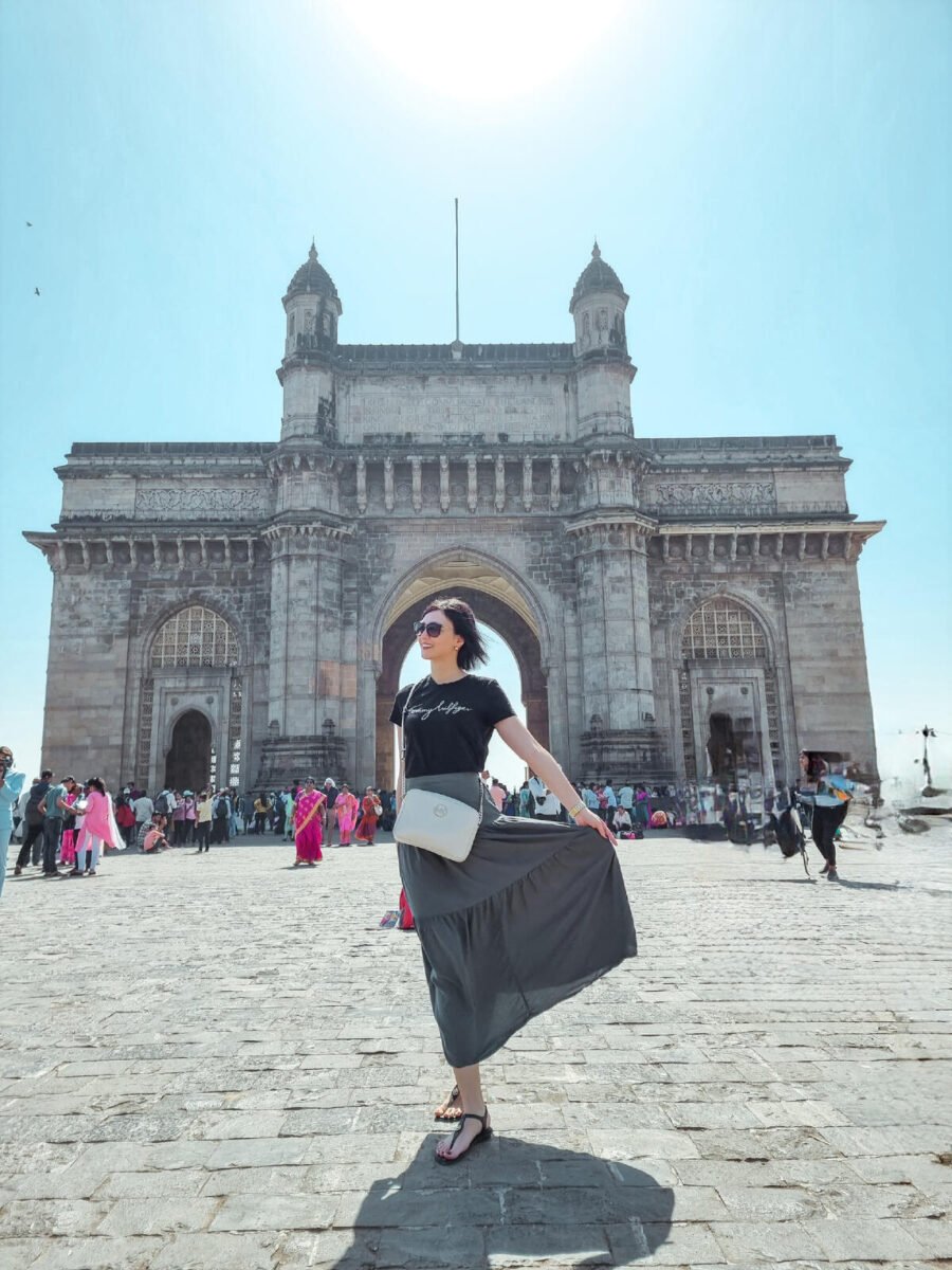 Female tourist posing in front of the iconic Gateway of India in Mumbai, with its grand arch and intricate colonial-era architecture surrounded by visitors on a bright sunny day.