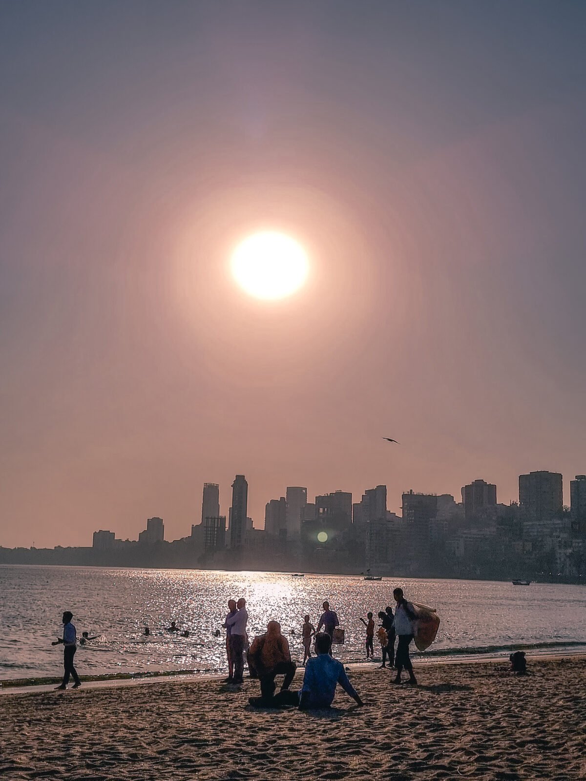 Silhouettes of people relaxing and walking along Girgaon Chowpatty Beach in Mumbai during sunset, with the shimmering Arabian Sea and the city skyline in the background.