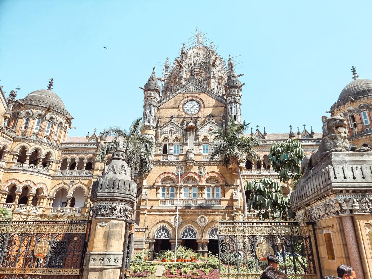 Front view of Chhatrapati Shivaji Maharaj Terminus in Mumbai, showcasing its stunning Victorian Gothic architecture with intricate details, arched windows, and a prominent clock tower surrounded by lush greenery.