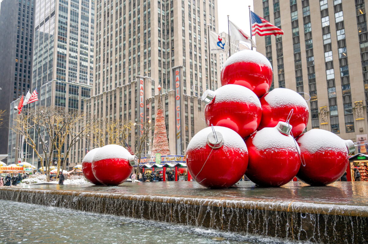 A festive winter scene in Midtown Manhattan featuring a large fountain on Sixth Avenue with a stack of oversized, bright red Christmas ornaments partially covered in fresh snow. In the background, the iconic Radio City Music Hall neon sign and a towering, lit-up Christmas tree are visible against the backdrop of soaring limestone skyscrapers. American and corporate flags flutter above the snowy street, which is lined with bare trees decorated with twinkling fairy lights.