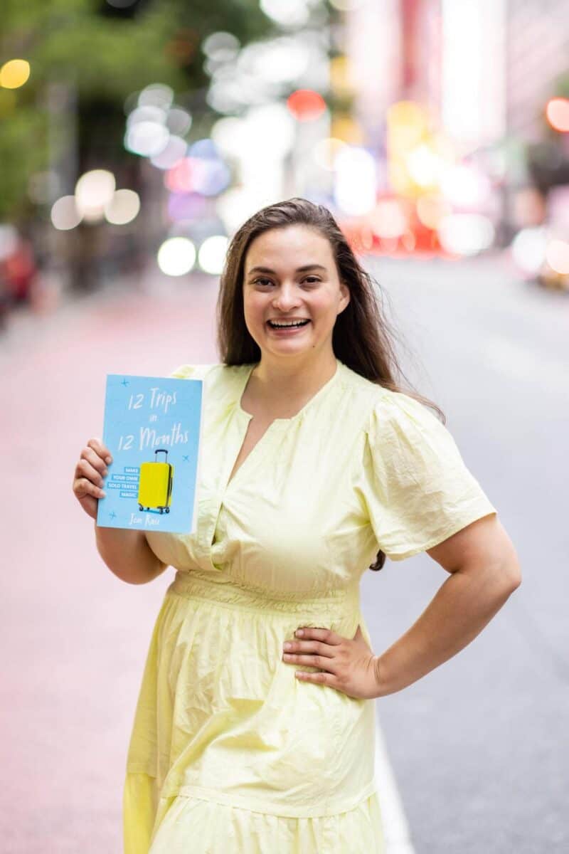 author Jen Ruiz wearing a yellow dress and holding up a copy of her published travel memoir