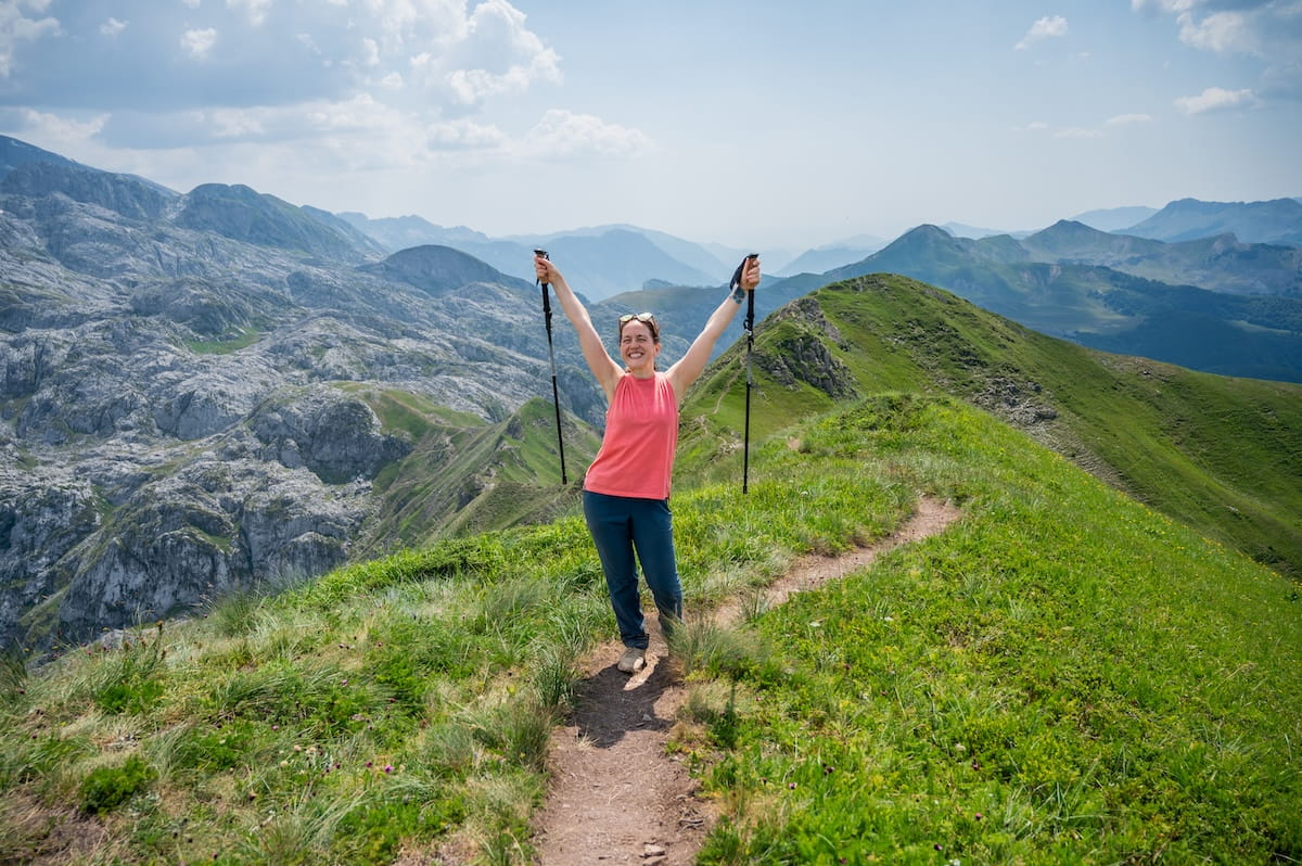 A woman in a pink tank top and blue hiking pants stands on a narrow mountain ridge with her arms raised victoriously, holding trekking poles. She is smiling at the camera against a backdrop of the dramatic, jagged gray and green peaks of the Peaks of the Balkans trail under a bright, partly cloudy sky.
