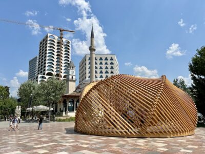 Modern wooden pavilion structure in Skanderbeg Square, Tirana, Albania, with the Et'hem Bey Mosque, clock tower, and modern high-rise buildings in the background on a sunny day.
