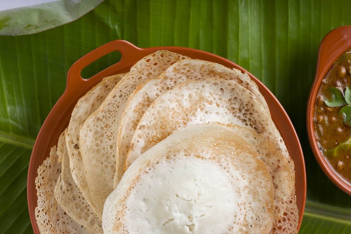 Close-up of freshly cooked South Indian appamโsoft, lacy-edged rice and coconut milk pancakesโserved in a brown dish on a banana leaf with curry on the side.