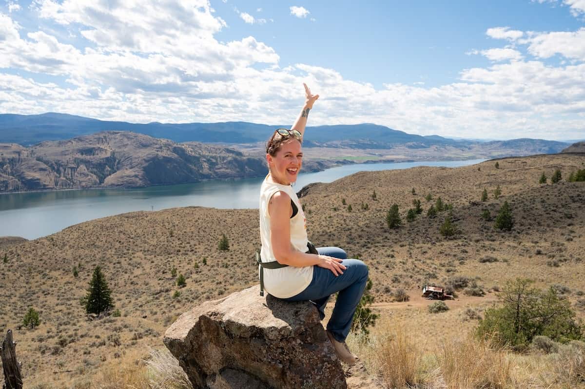 Woman on a British Columbia road trip sitting on a rock and smiling while pointing toward the scenic view from Battle Bluff Trail in Kamloops, with dry rolling hills, scattered pine trees, and Kamloops Lake shimmering under a bright blue sky.