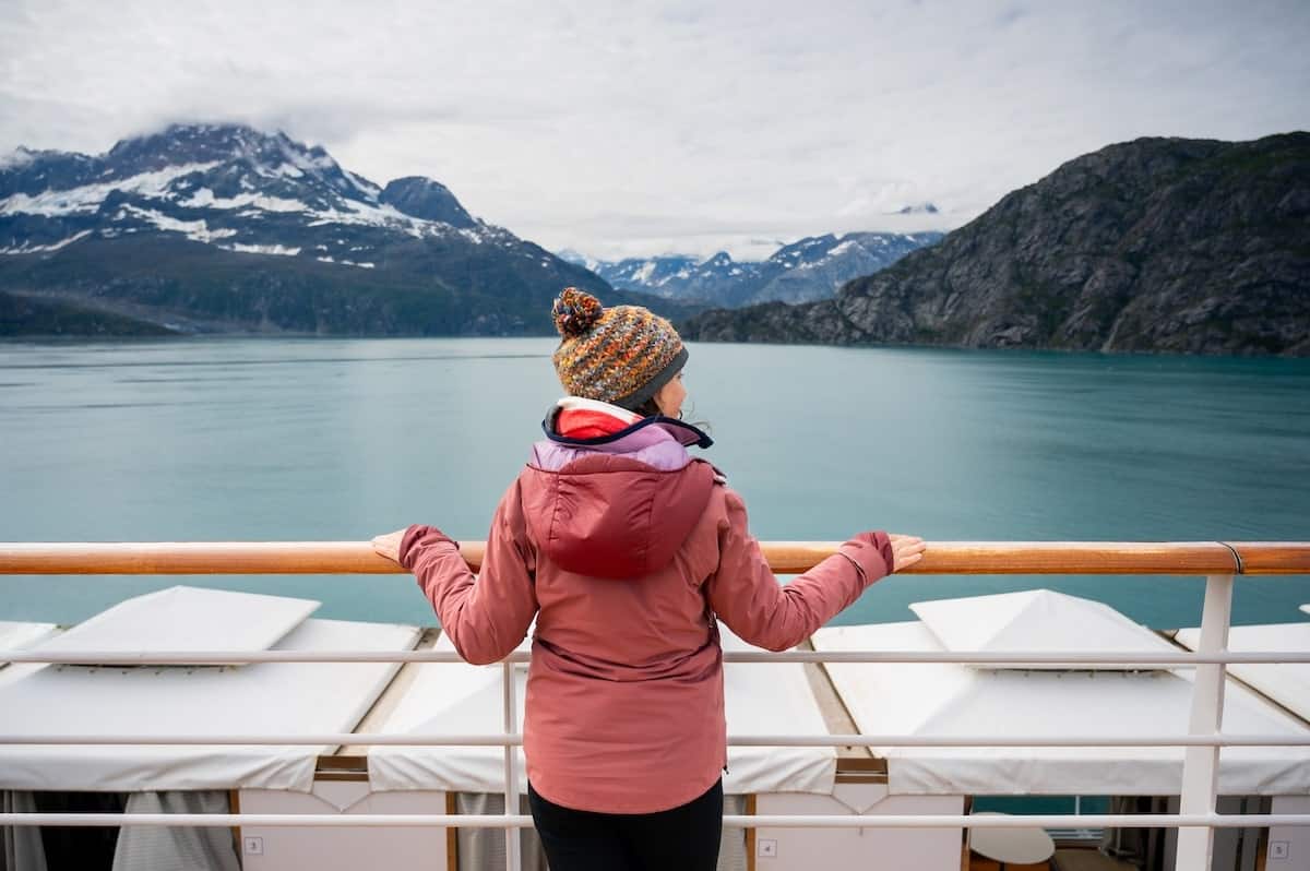 Traveler in a pink jacket and knit hat admiring the calm turquoise waters and dramatic mountain peaks of Glacier Bay National Park from the deck of a Holland America Alaska cruise ship.