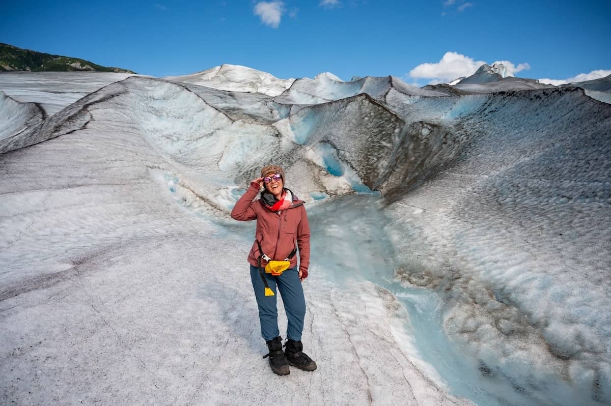 Traveler standing on Meade Glacier near Skagway, Alaska, smiling and holding sunglasses on a guided helicopter tour, surrounded by blue ice formations and rugged glacier terrain under a clear sky.