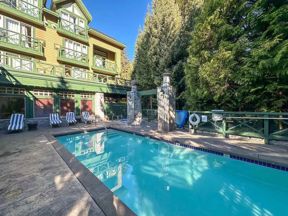 Outdoor pool at the Pinnacle Hotel Whistler Village, surrounded by striped lounge chairs, forest trees, and the hotelโs green-and-yellow exterior.