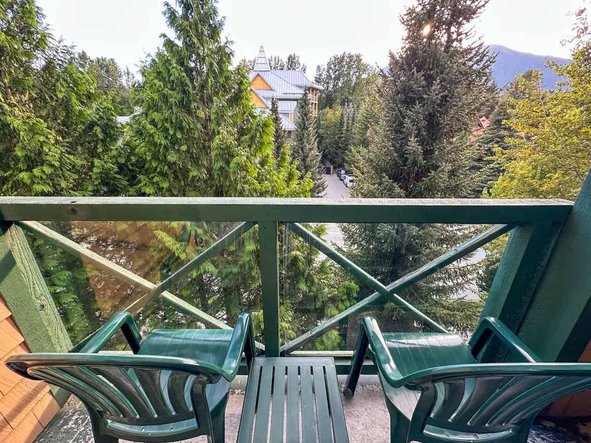 Private balcony at Pinnacle Hotel Whistler Village with two green chairs and a small table overlooking tall evergreens and distant mountains on a clear day.
