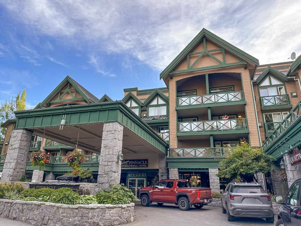 Front exterior of the Pinnacle Hotel Whistler Village with green trim and stone pillars, showing the covered entrance, hanging flower baskets, parked cars, and guest balconies against a partly cloudy blue sky.