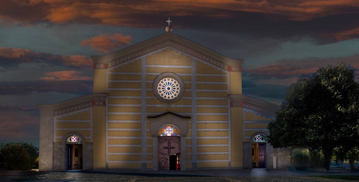 A frontal view of the Shkodër Cathedral (St. Stephen’s Cathedral) at dusk, illuminated by warm exterior lighting. The church features a symmetrical neoclassical design with horizontal white stripes across a yellow façade, three arched doorways with stained glass windows above, and a circular rose window in the center. The sky behind is painted with dramatic purples and oranges, and a large tree partially frames the right side of the scene.