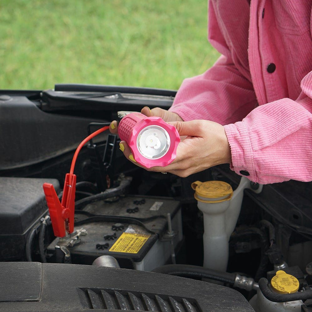 A person wearing a pink shirt holds a bright pink Limitless BoostHero portable jump starter and flashlight over an open car engine bay. The deviceโs LED lens is pointed toward the camera. In the background, a red jumper cable clamp is attached to the car battery terminal. The scene is set outdoors against a soft-focus green grassy background.