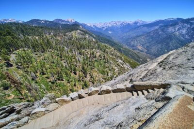 View looking down the steep stone staircase of the Moro Rock Trail in Sequoia National Park, California, with rugged granite cliffs and sweeping views of forested mountains and valleys in the distance under a clear blue sky.