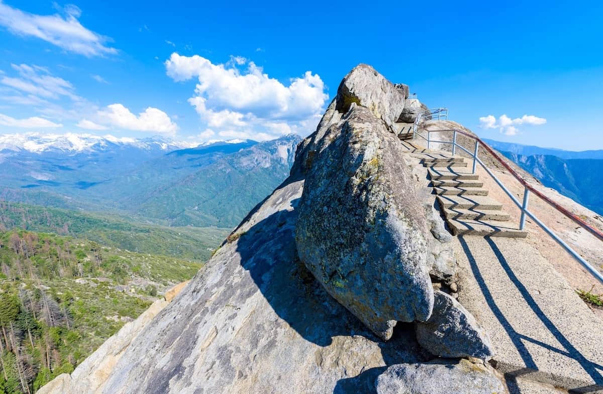 Steep staircase with railings leading up the granite summit of Moro Rock in Sequoia National Park, California, overlooking snow-capped Sierra Nevada peaks and lush green valleys under a bright blue sky.