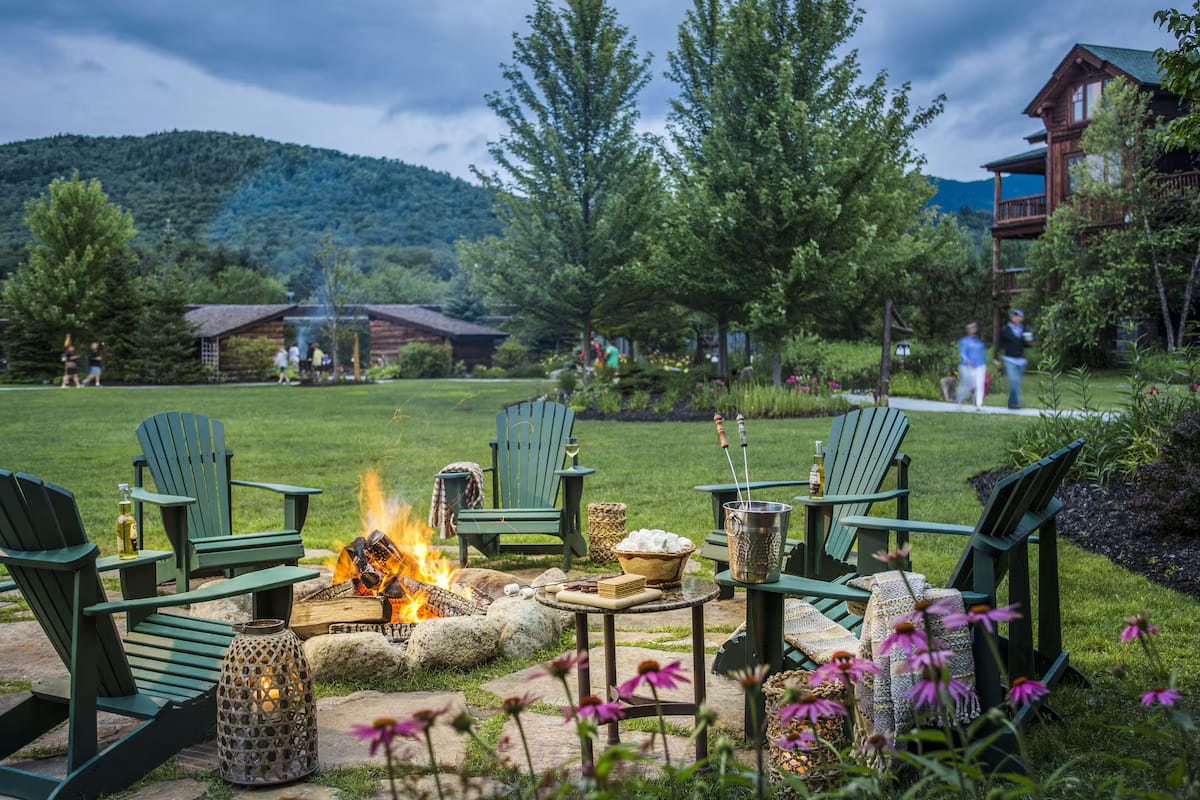 An outdoor evening patio scene on the grounds of The Whiteface Lodge in Lake Placid, New York, a top Thanksgiving getaway. It features a stone fire pit burning with bright flames surrounded by green Adirondack chairs on a grassy lawn. On a small stone side table sit s'mores ingredients including marshmallows and graham crackers, alongside a silver ice bucket and drink bottles. Pink coneflowers frame the bottom edge, with towering pine trees, rustic timber lodge buildings, and lush green mountains under a cloudy dusk sky in the background.