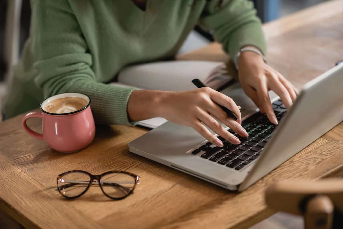 Female blogger typing up her substack newsletter. She is wearing a green sweater and holding a blank pen while her fingers touch the laptop keys.