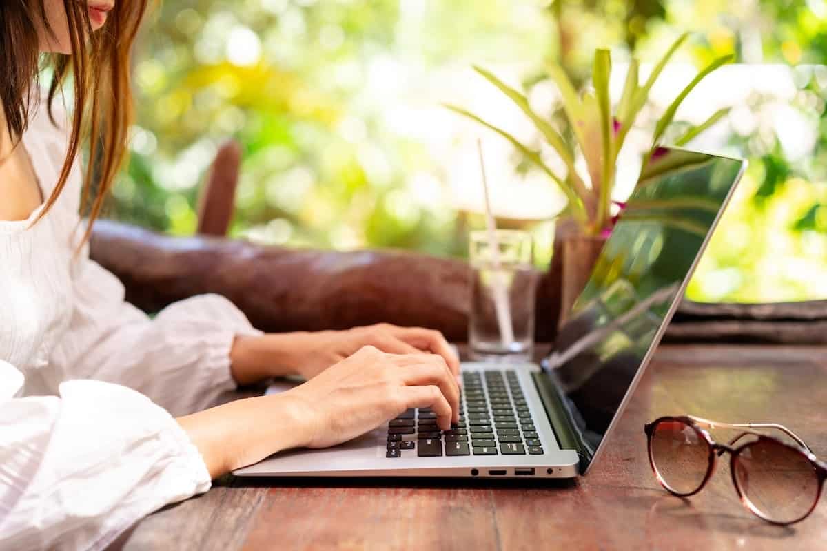 A woman works on her laptop outdoors while writing a travel newsletter, with sunglasses and a glass of water on a wooden table surrounded by tropical greenery.