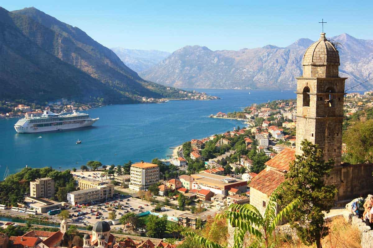 A sweeping view of Kotor Bay featuring a large cruise ship sailing through deep blue water, a coastal town with red-tiled roofs below, and dramatic mountain slopes rising around the bay, with a stone church tower in the foreground.