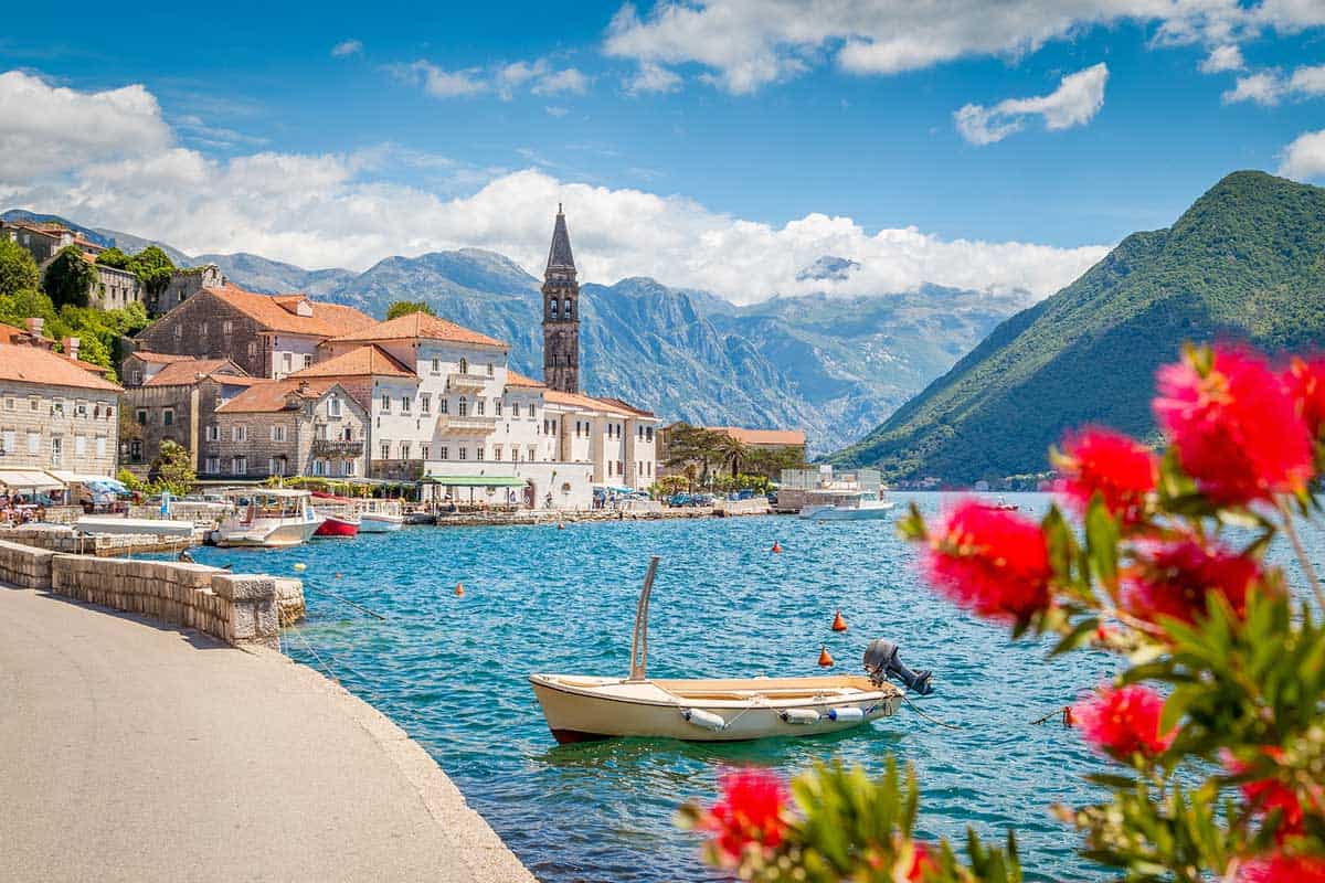 A picturesque seaside town with stone buildings and a church bell tower along the waterfront, small boats moored in turquoise water, mountains in the distance, and red flowers in the foreground under a bright blue sky.