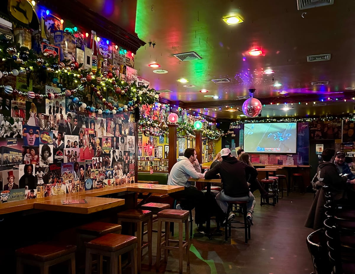 A wide shot of the interior of Somebodyโs Darling, a bar on NYC's Upper East Side featuring a vibrant retro-maximalist vibe with walls covered in pop culture collages, festive multi-colored string lights, a disco ball, and patrons seated at high-top wooden tables.