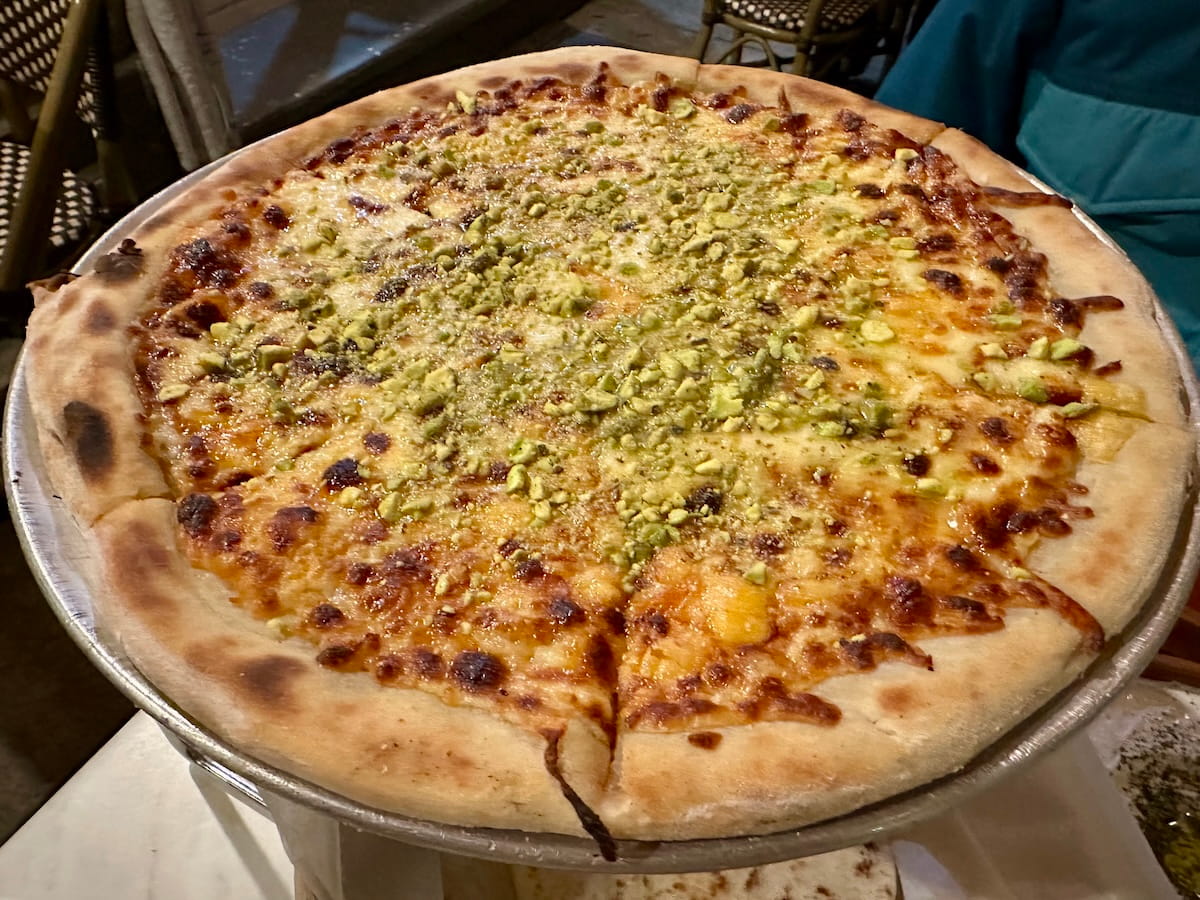 A large, round Pistachio & Cheese Pizza served on a metal tray at Al Badawi Palestinian Restaurant on NYC's Upper East Side. The pizza features a thick, golden-brown crust and is covered in a rich, melted cheese layer topped with a generous dusting of crushed bright green pistachios.