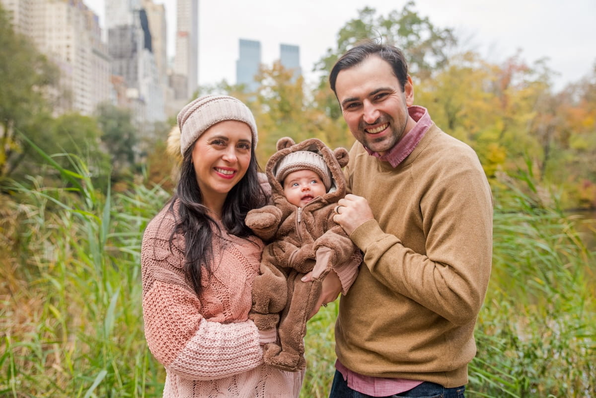 A medium, eye-level shot of a smiling family of three posing outdoors in Central Park during autumn. A woman on the left wears a pink cable-knit sweater and a tan beanie, while a man on the right wears a tan crewneck sweater over a maroon collared shirt. They are holding a baby dressed in a fuzzy brown bear-ear hooded jumpsuit. The background features tall green and yellow marsh grasses, fall foliage, and the distant skyline of Central Park South under an overcast sky.