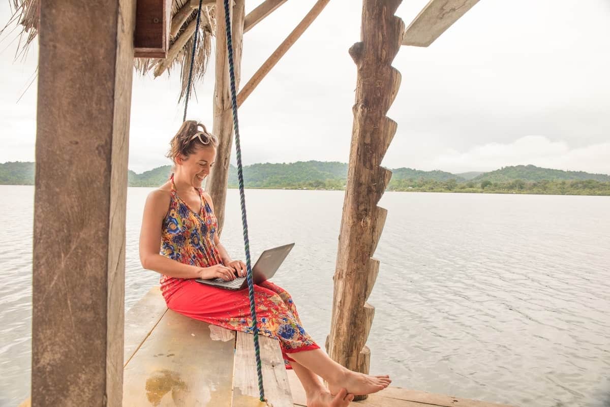 Travel blogger working on a laptop at a lakeside wooden dock, smiling while creating content surrounded by nature.