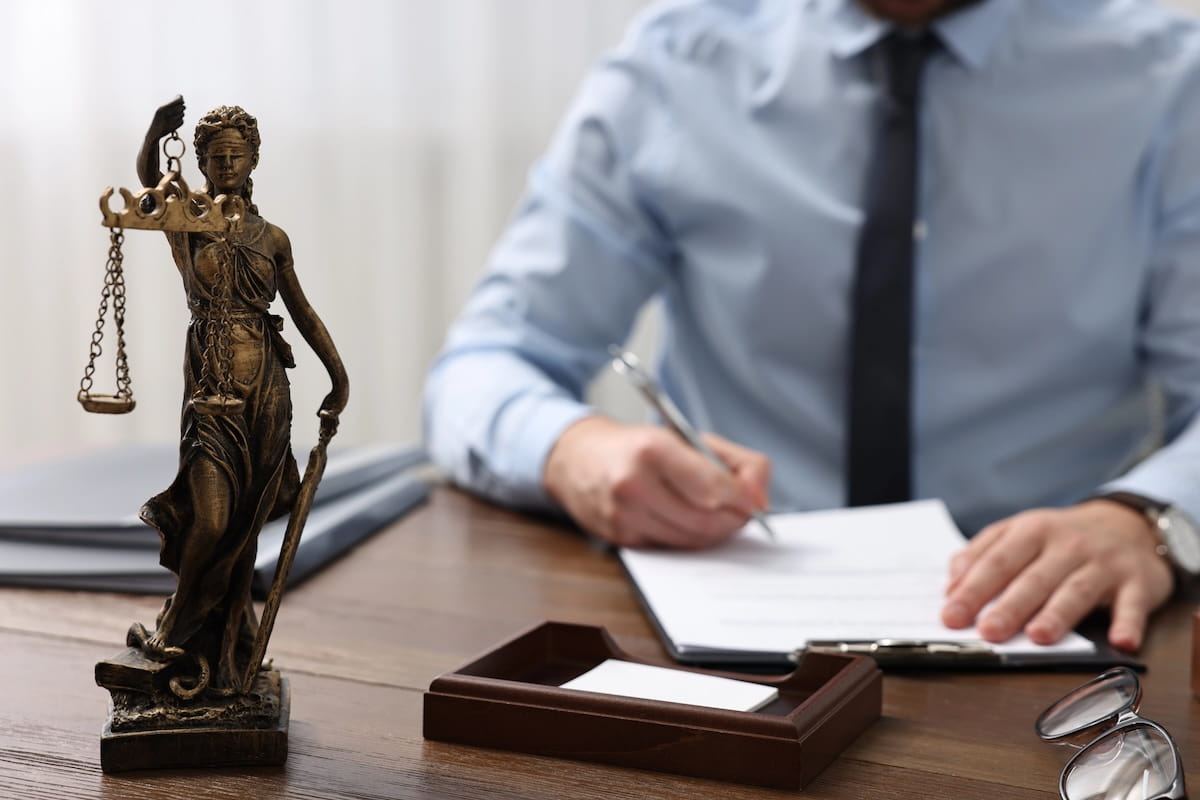 A legal statue of Lady Justice in the foreground with a lawyer in a light blue shirt and tie blurred in the background, writing on a document. This illustrates how the Copymight Coalition provides legal protection and compensation for bloggers against AI piracy.