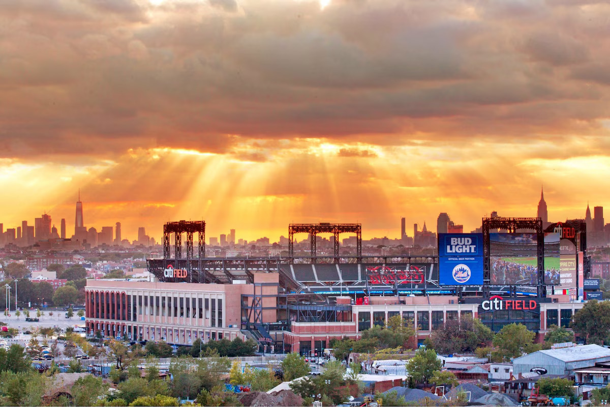 A wide-angle, high-angle landscape shot taken from the Compass rooftop at sunset overlooking Citi Field in Queens. Dramatic golden sunbeams break through a thick, dramatic cloud cover, illuminating the entire sky in warm orange hues. In the midground, the exterior architecture and large jumbotron of Citi Field stadium are fully visible. The Manhattan skyline—featuring One World Trade Center on the far left and the Empire State Building on the right—stretches along the horizon beneath the glowing sunset.