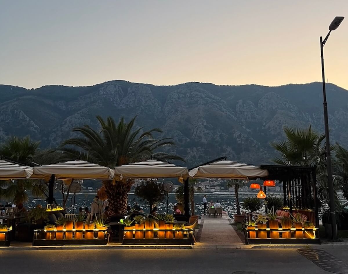 An outdoor waterfront restaurant terrace at dusk, with striped umbrellas, warm candlelit planters, and palm trees, backed by dramatic mountain silhouettes and a marina in the Bay of Kotor, Montenegro.