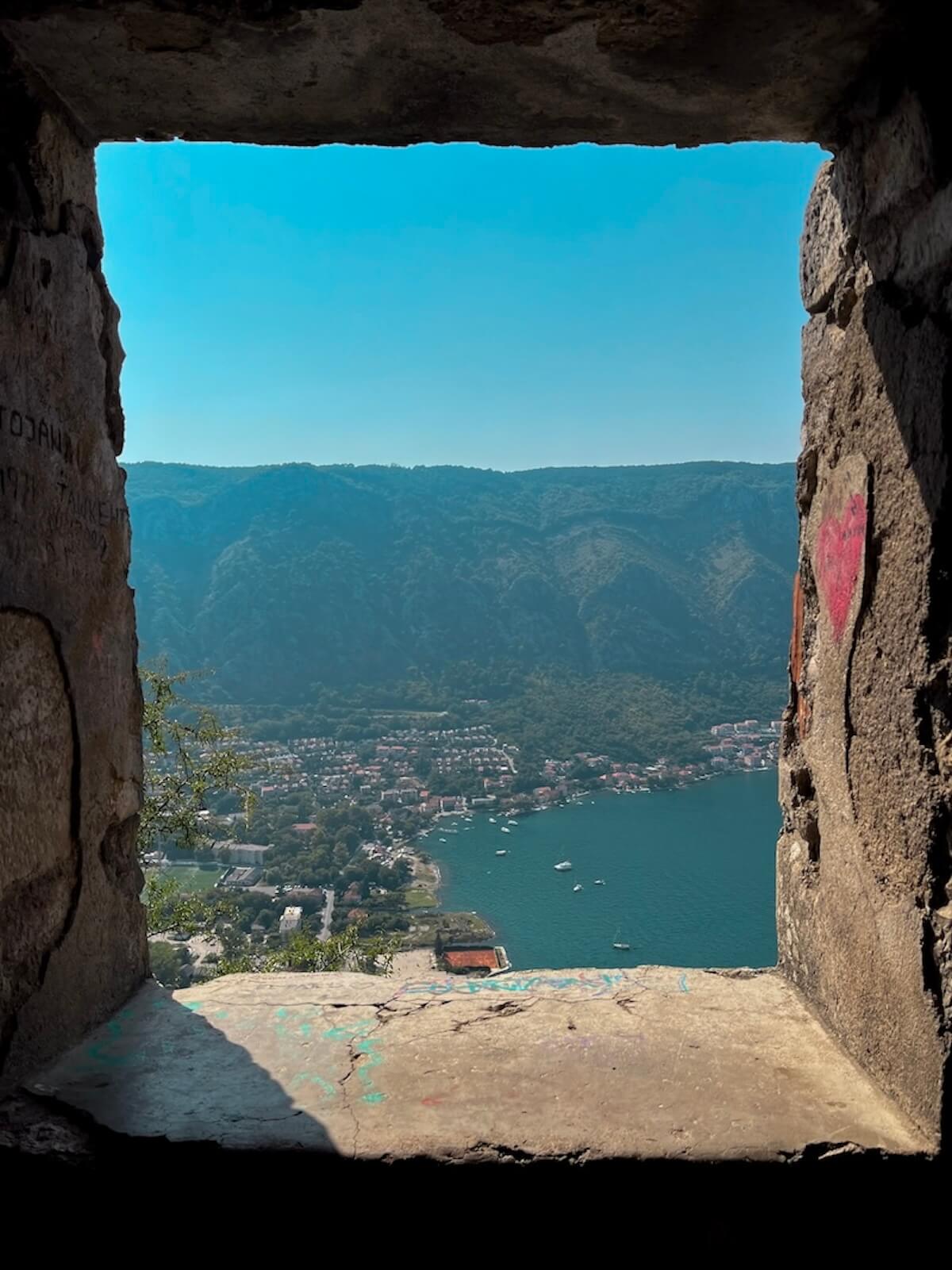 A view of the Bay of Kotor and a hillside town framed through a rectangular stone window opening in Kotor's ancient fortress walls, with green mountains and turquoise water below.