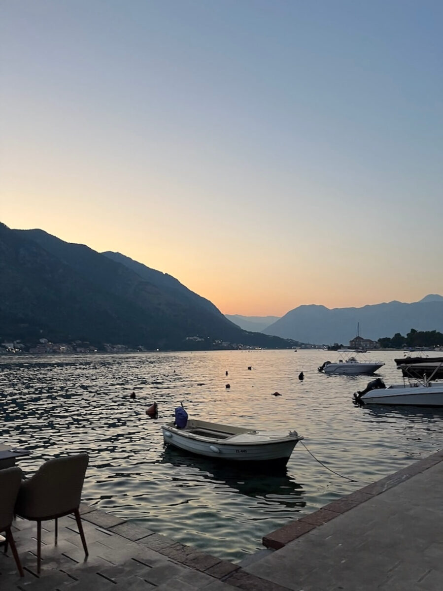 A small white rowboat moored at a stone waterfront promenade at sunset, with calm water reflecting pastel skies and layered mountain silhouettes in the background, Perast, Montenegro.
