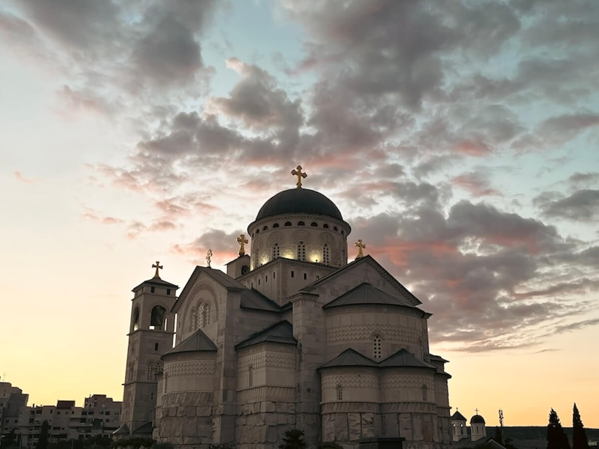 A large Serbian Orthodox cathedral with a dark dome and golden crosses photographed from below at sunset, with dramatic pink and grey clouds illuminated in the sky behind it, Podgorica, Montenegro.