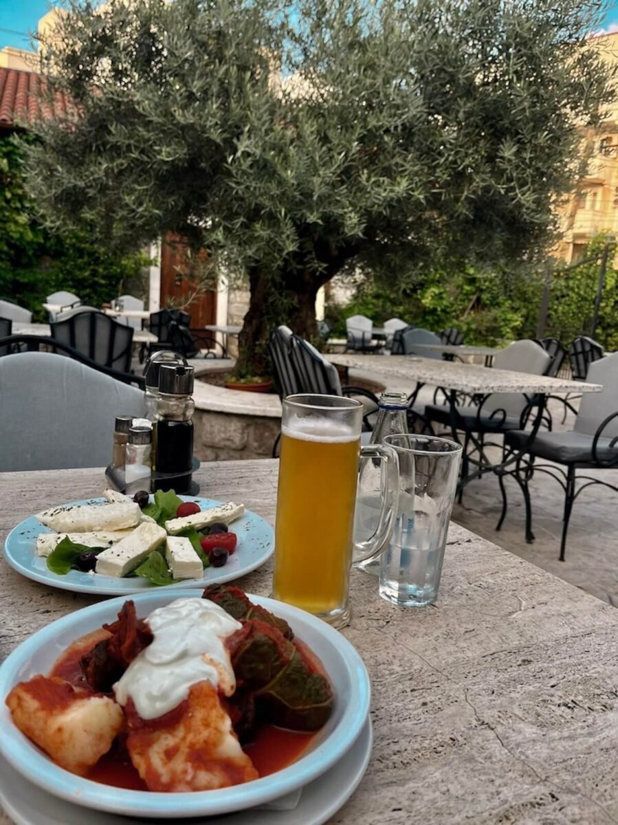 An outdoor restaurant table beneath a large olive tree in Montenegro, set with a plate of white cheese salad, a bowl of stuffed grape leaves topped with sour cream, and a tall glass of draft beer.