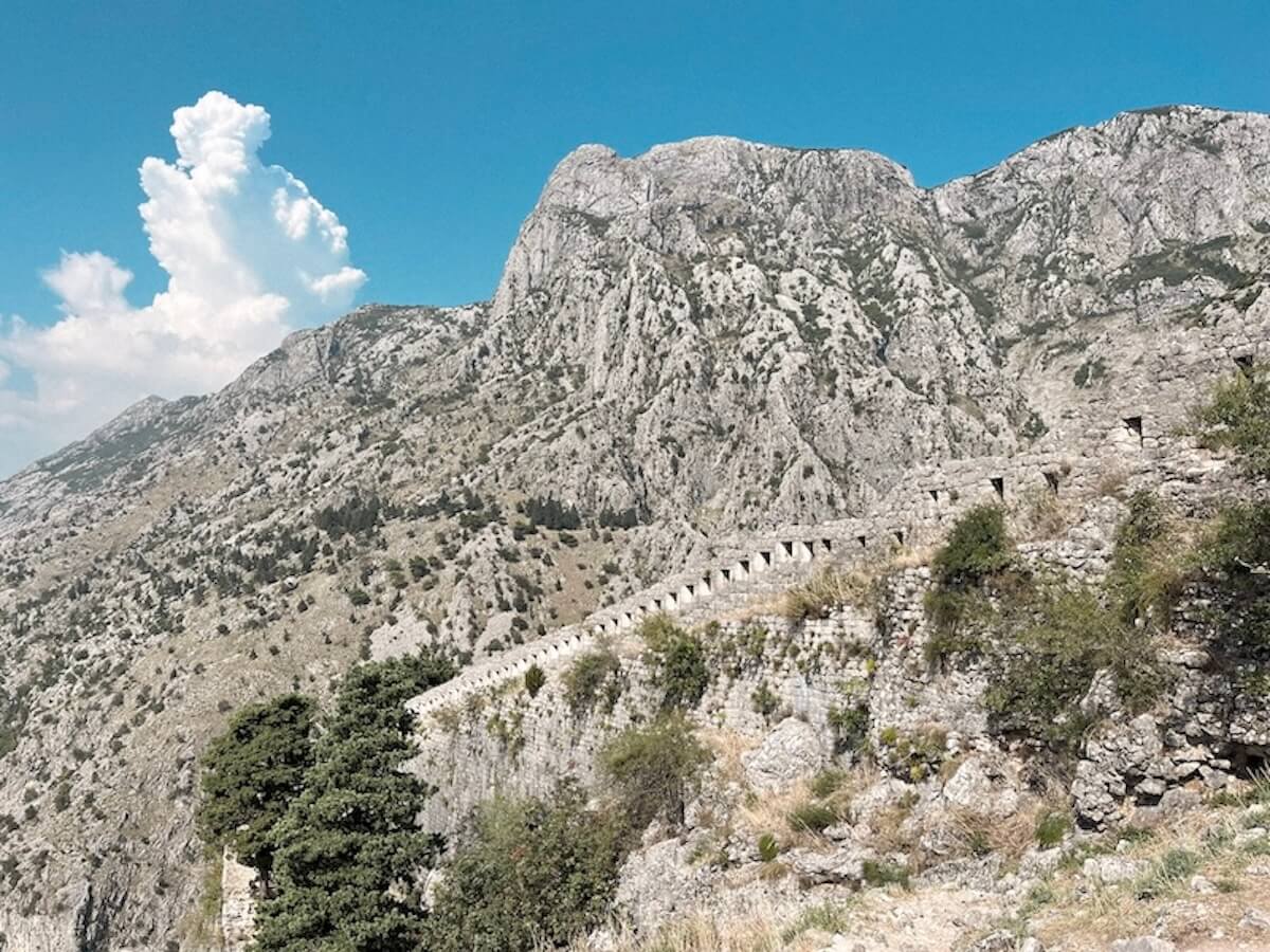 The ancient serpentine stone defensive walls of Kotor's fortress climbing steeply up a dramatic limestone mountain face under a bright blue sky, Montenegro.