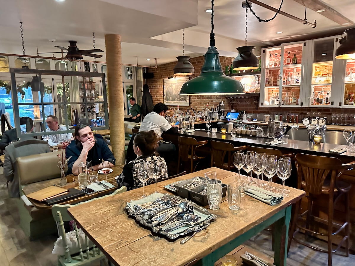 The cozy, rustic interior of Chez Rioux & Pettigrew restaurant in Quebec City, featuring weathered wood tables, exposed brick walls, and vintage decor. A central wooden table is neatly set with silver platters of cutlery and wine glasses, while patrons dine at a dark bar under industrial green pendant lights.