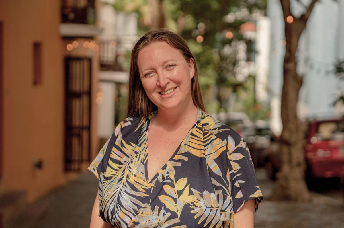 Travel creator and freelancer Brittany Verlenich smiling in a tropical outdoor setting with blurred buildings and trees in the background.