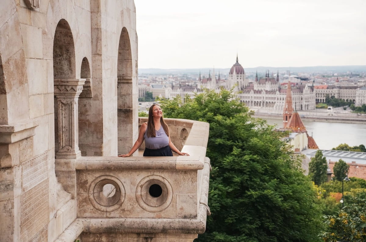 Female freelancer smiling on a balcony in Budapest with the Hungarian Parliament building in the background, illustrating a high-value freelance service idea.